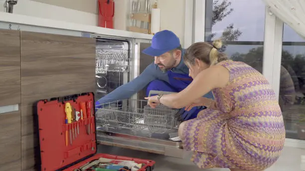 Technician repairing a dishwasher with a homeowner, showcasing appliance repair services in a modern kitchen setting.