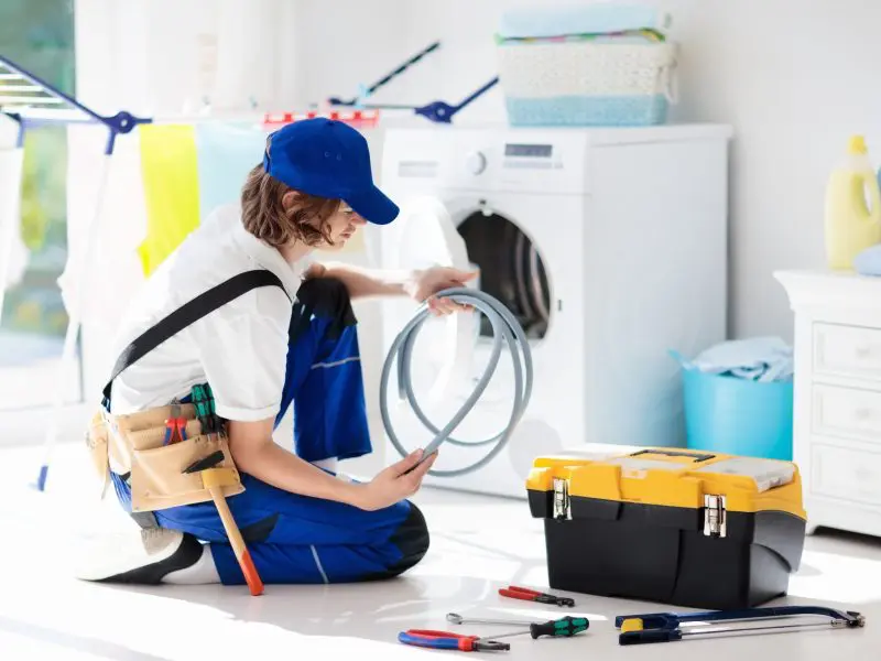 Technician in blue uniform and cap examining washing machine hose, surrounded by tools and laundry equipment, illustrating professional appliance repair service.