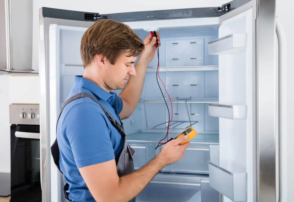 Technician repairing Insignia refrigerator using multimeter for diagnostics in modern kitchen setting.